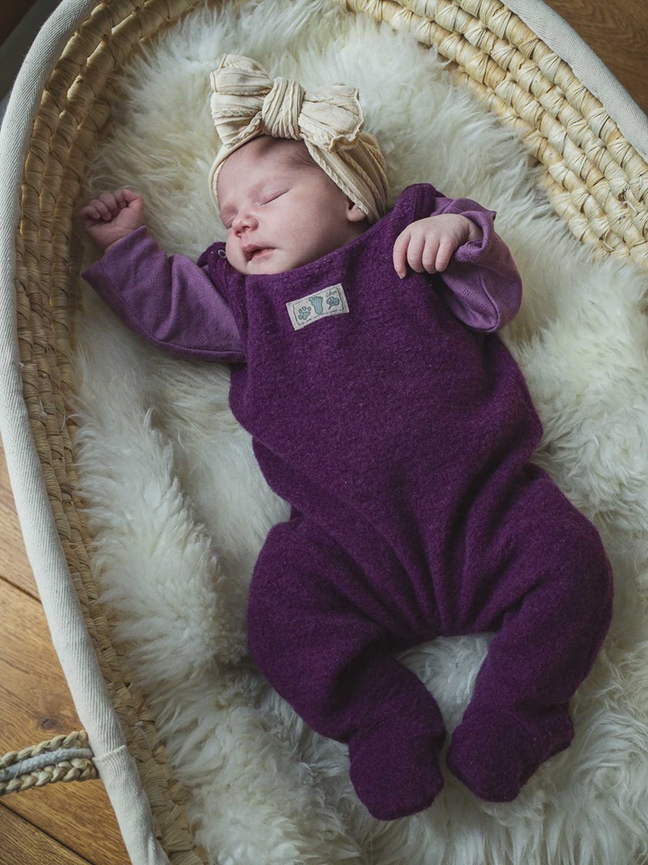 Ein schlafendes Baby in lila Wollanzug mit beigem Haarband in einem Korb auf einem weichen Schaffell. Newbornshooting in und um Geretsried