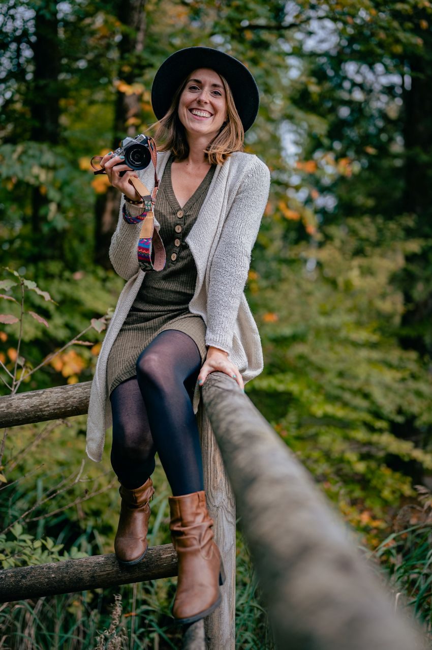 Junge Frau mit Kamera in der Hand, sitzt lächelnd auf einem Baumstamm im Wald.