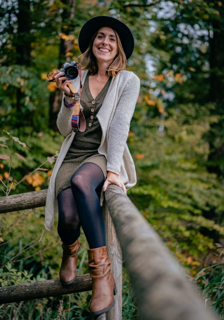 Frau mit Hut sitzt auf einem Holzzaun, hält eine Kamera und lächelt, im Hintergrund ein herbstlicher Wald. Fotografie in Geretsried.