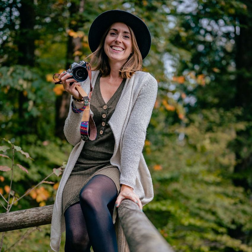 Lächelnde Frau mit Kamera und Hut in einer herbstlichen Waldumgebung.
