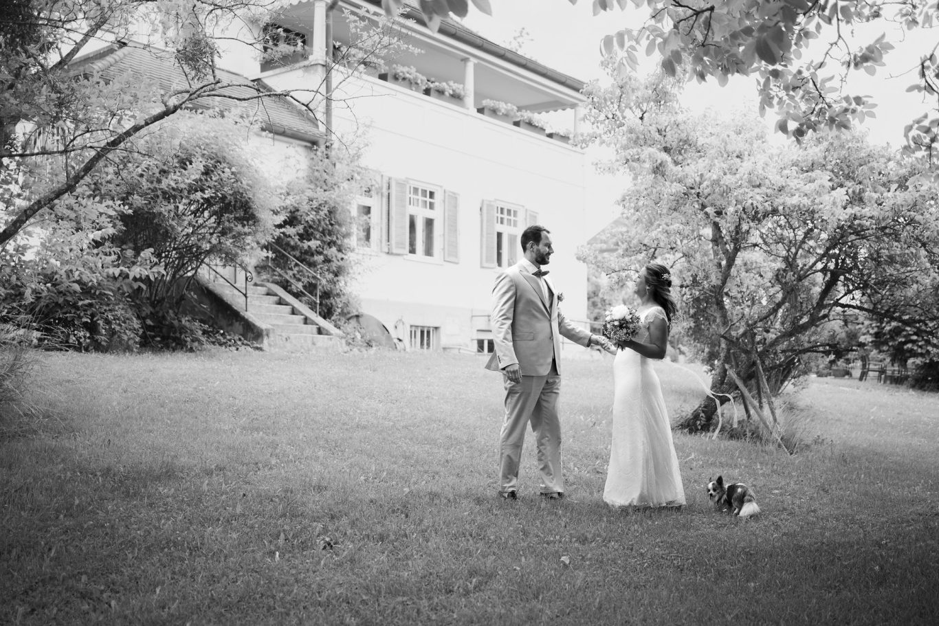 Hochzeit schwarz-weiß-Fotos des Brautpaares wie es vor einer Villa auf einer Wiese zwischen Bäumen steht und sich einander glücklich ansieht
