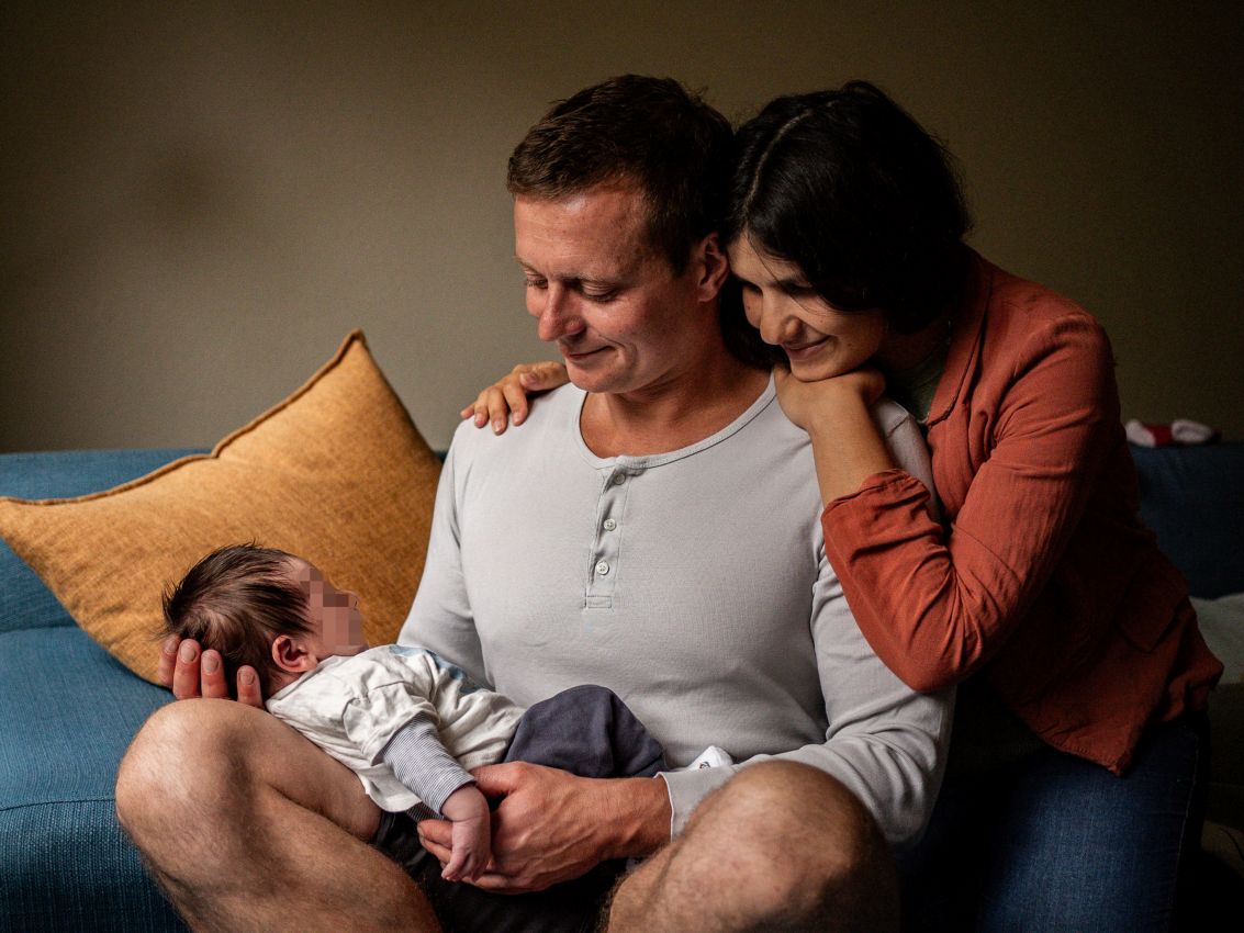 Ein glückliches Paar sitzt mit ihrem Neugeborenen auf dem Schoß auf einer Couch und lächelt es an. Fotografie in Geretsried, Wolfratshausen.