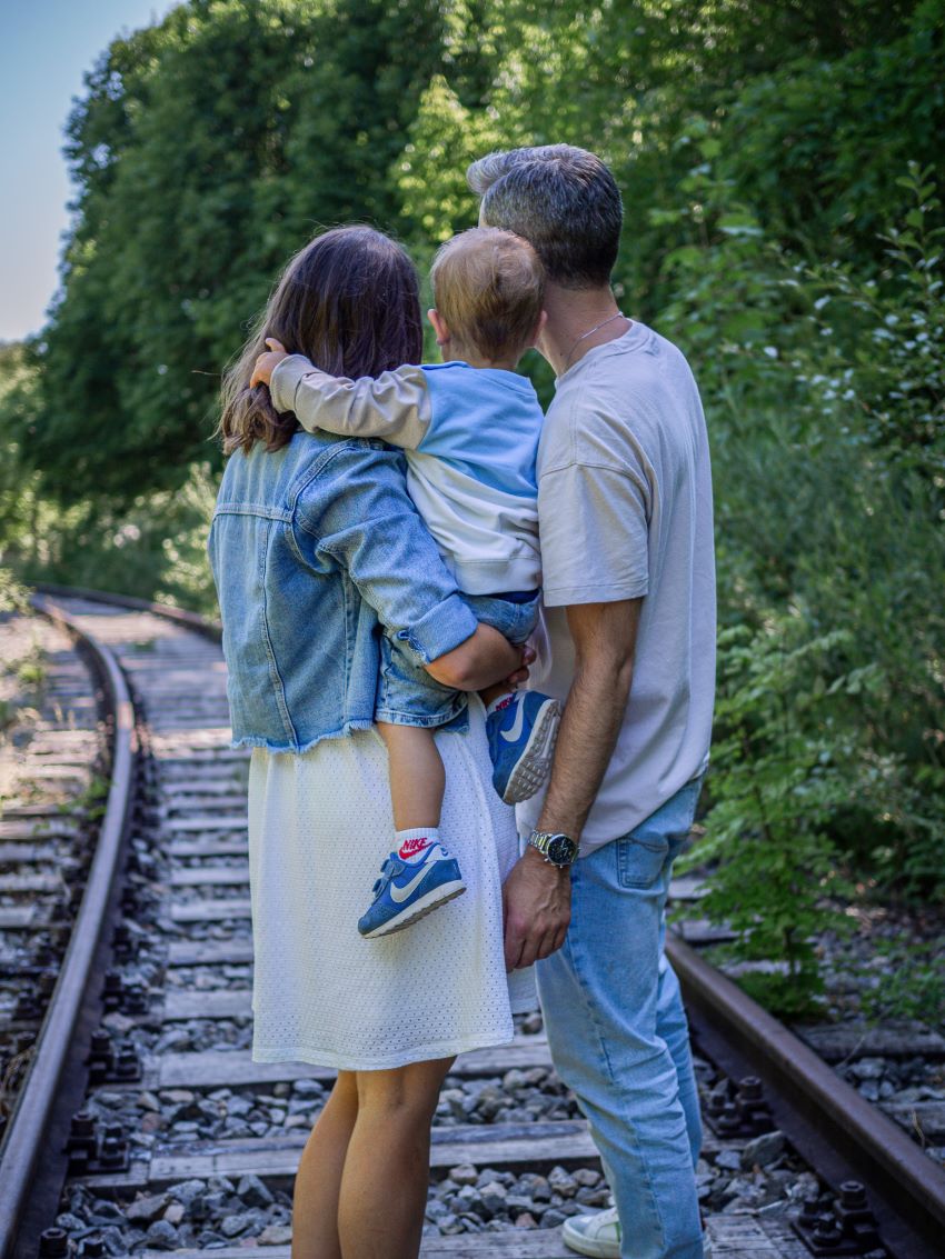 Familie steht an einer alten Gleisanlage und blickt in die Natur, das Kind wird in der Mitte von beiden Eltern gehalten. 