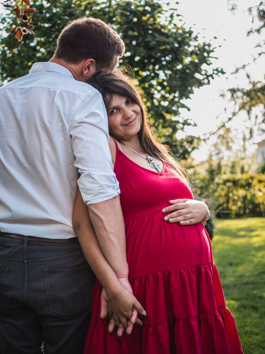 Schwangere Frau in rotem Kleid, lehnt sich an die Schulter ihres Mannes im weißen Hemd, in einem Garten, ihre Hand auf dem runden Bauch.