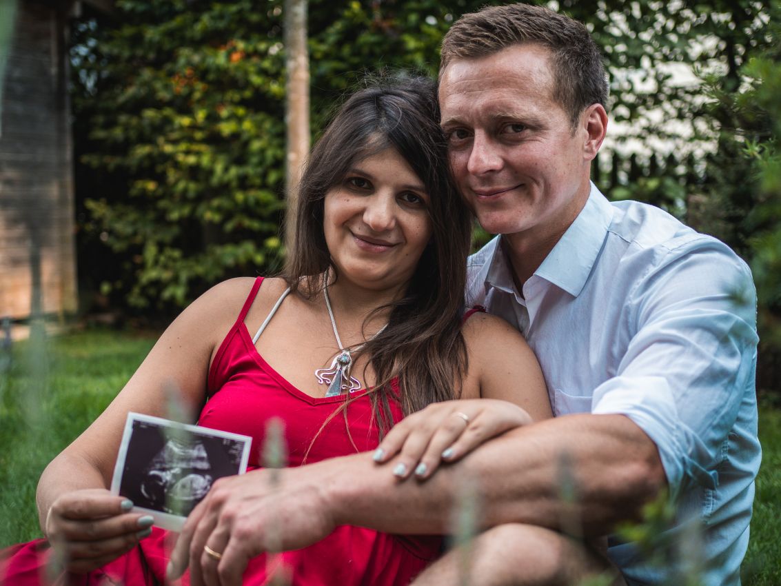 Paar sitzt Arm in Arm in einem Garten und hält ein Ultraschallbild in der Hand. Babybauchshooting in und um Geretsried Wolfratshausen