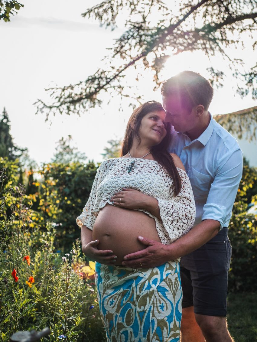 Ein Paar umarmt sich während des Sonnenuntergangs liebevoll auf einer Wiese, während sie den Babybauch der schwangeren Frau streicheln.