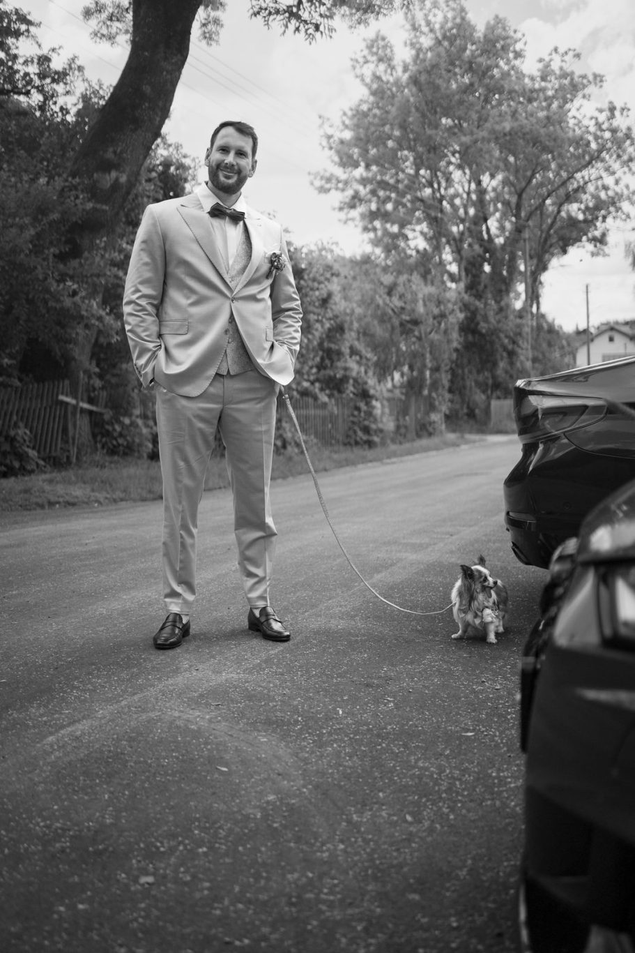 Hochzeit schwarz-weiß-Foto des Bräutigams, wie er mit seinem kleinen Hund an der Leine auf der Straße steht und zufrieden lächelt