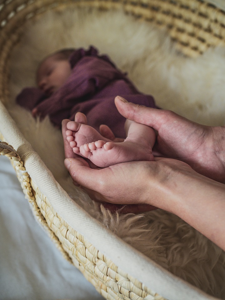 Neugeborenes Baby, das in einem Korb liegt, während zwei Hände die Füße des Babys halten. Newbornshooting in und um Geretsried