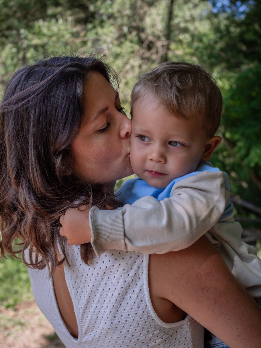 Eine Mutter küsst ihr kleines Kind, beide lächeln in einer natürlichen Umgebung.
