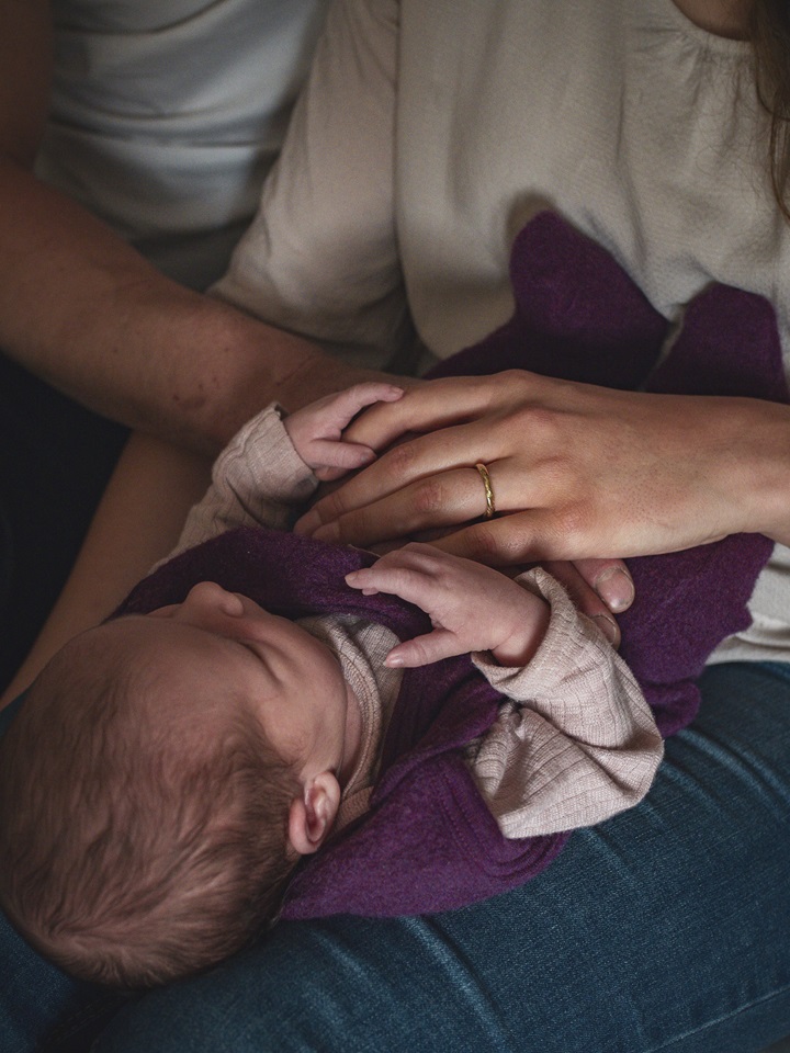 Neugeborenes Baby in lila Strampler, liegt auf dem Schoß der Mutter. Die Eltern legen liebevoll ihre Hände auf den Bauch des Babys. 