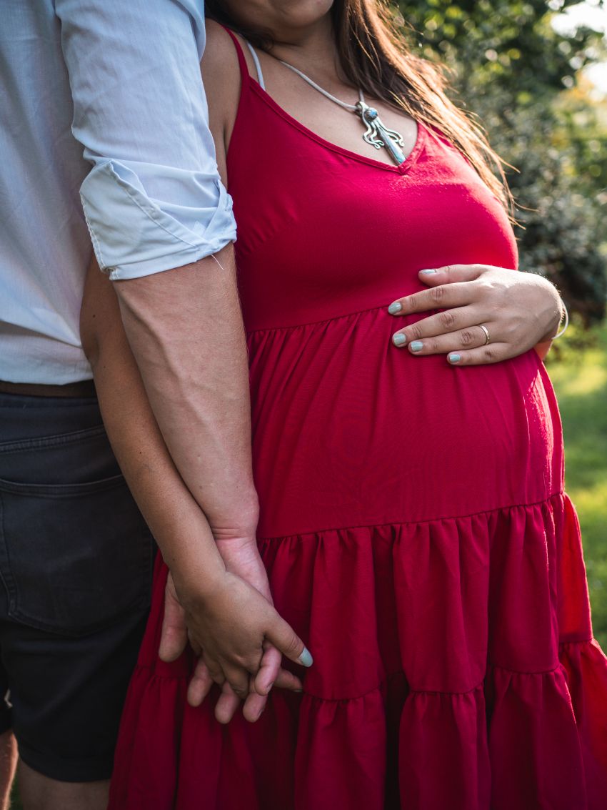 Schwangere Frau in rotem Kleid hält die Hand ihres Partners im Freien. Babybauchshooting in und um Geretsried Bad-Tölz Wolfratshausen