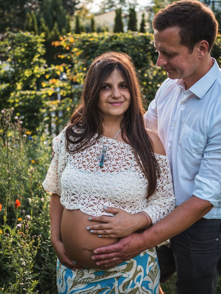 Schwangere Frau in einem Garten, lächelt in die Kamera und hält ihren Bauch, ihr Mann umarmt sie liebevoll. 