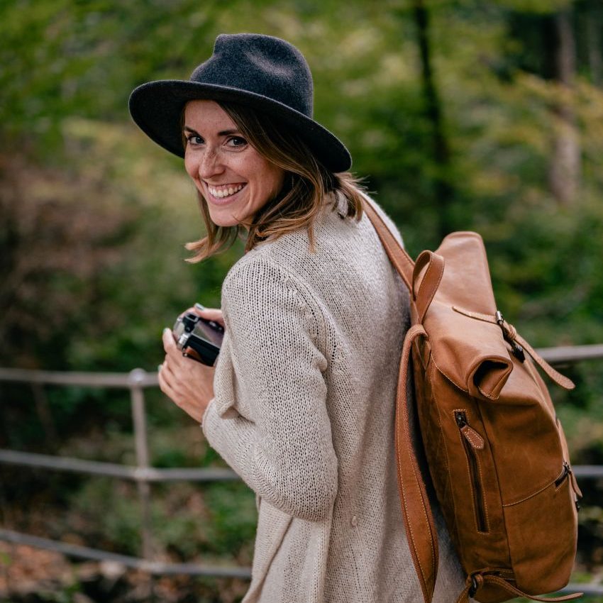 Junge Frau mit Hut und Rucksack, die lächelnd eine Kamera hält, vor einem Waldhintergrund.
