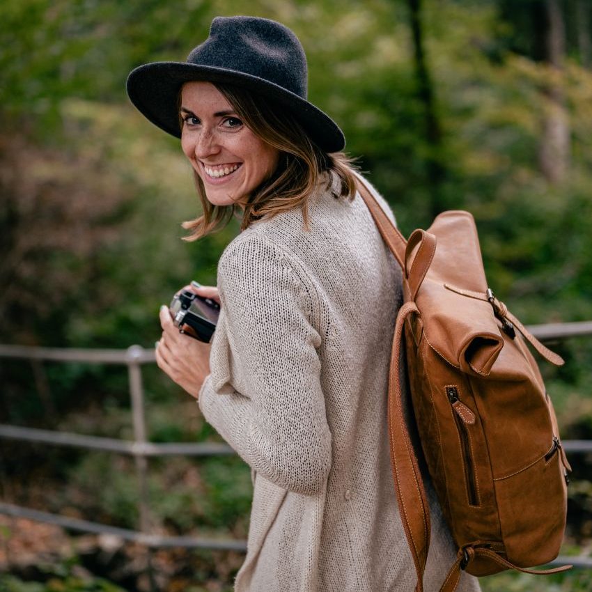 Frau mit Kamera, grauem Hut und braunem Rucksack, lächelnd im Wald.